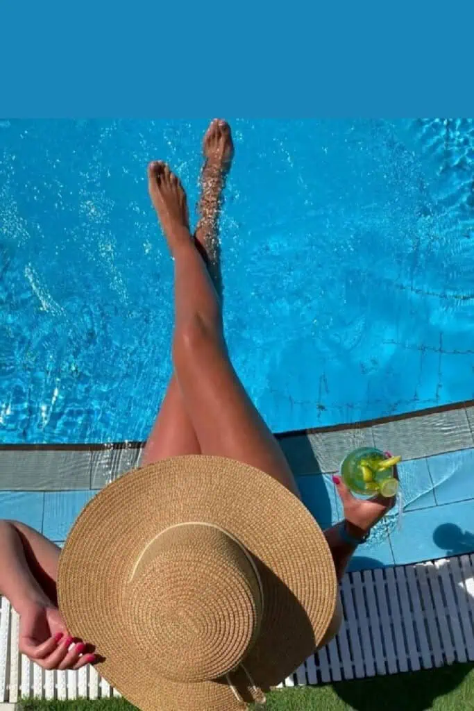Woman lounging poolside with legs dipped in the water, wearing a straw sunhat and holding a summer cocktail with lemon and mint—perfect summer aesthetic shot for vacation vibes and Instagram-worthy poolside relaxation.