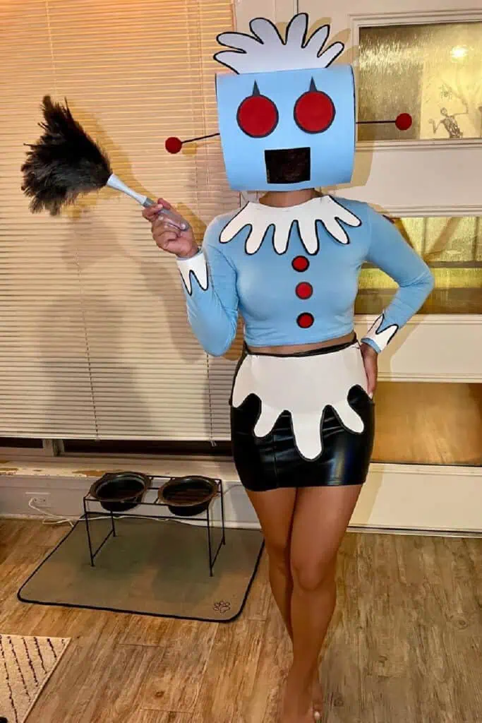 DIY Rosie the Robot costume inspired by The Jetsons. A creative handmade Halloween outfit featuring a light blue top with red buttons, white cartoon-style collar and apron, black skirt, and a box robot headpiece with red eyes. The person holds a feather duster to complete the look.