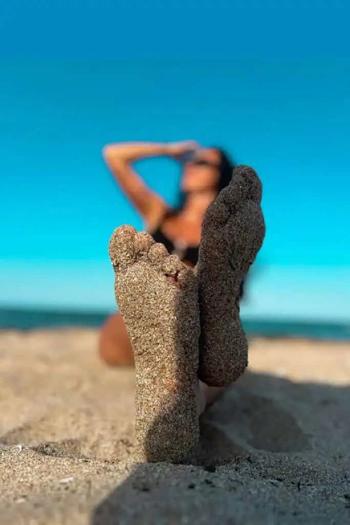 Sandy feet close-up with a woman lounging on the beach in the background, toes pointed at the camera under a clear blue sky—fun, relaxed summer photo with a dreamy vacation vibe.