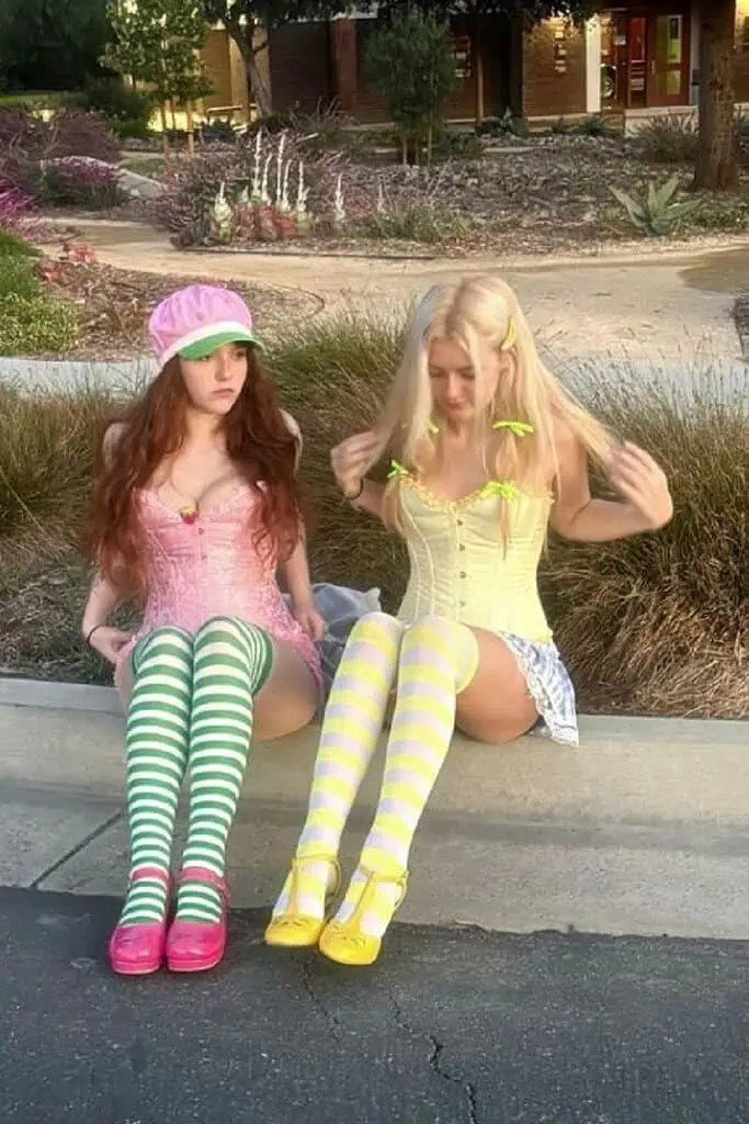 Two women in duo Halloween costumes dressed as Strawberry Shortcake and Lemon Meringue. Strawberry Shortcake wears a pink corset dress with a strawberry hat, green-and-white striped stockings, and pink shoes. Lemon Meringue wears a yellow corset dress with yellow-and-white striped stockings, yellow bows in her hair, and yellow shoes.