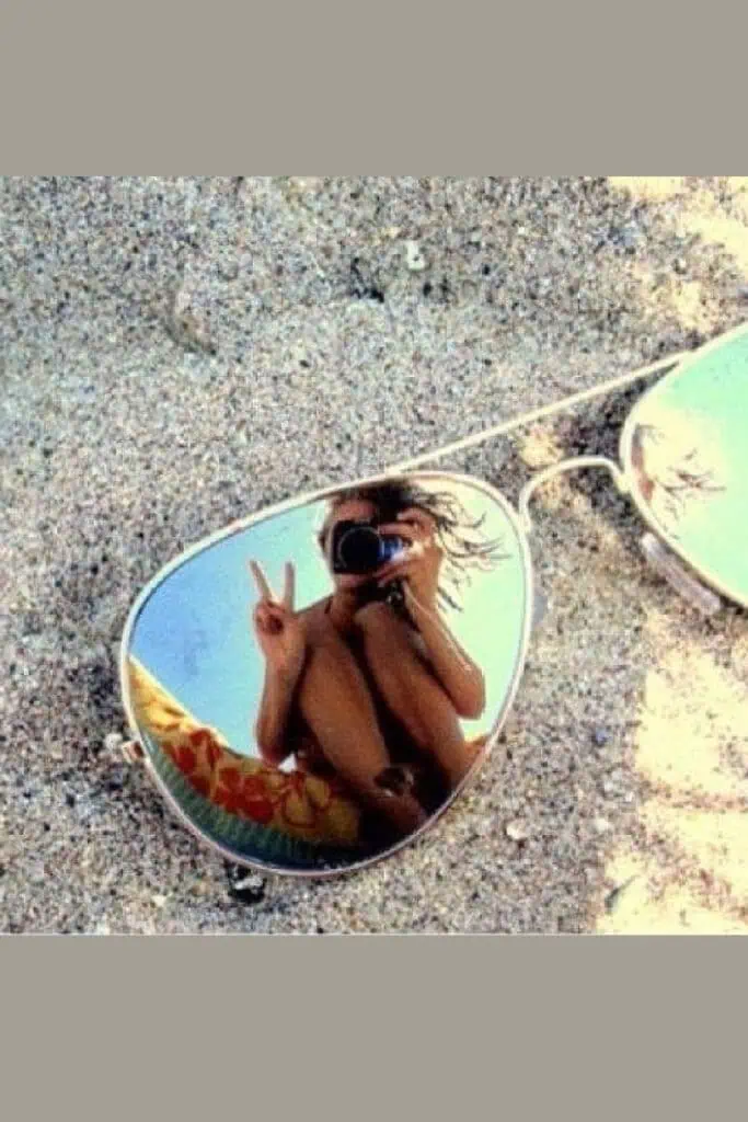 Creative beach selfie showing a woman’s reflection holding a camera and peace sign in mirrored aviator sunglasses lying on the sand—playful summer shot with a fun, artsy twist.