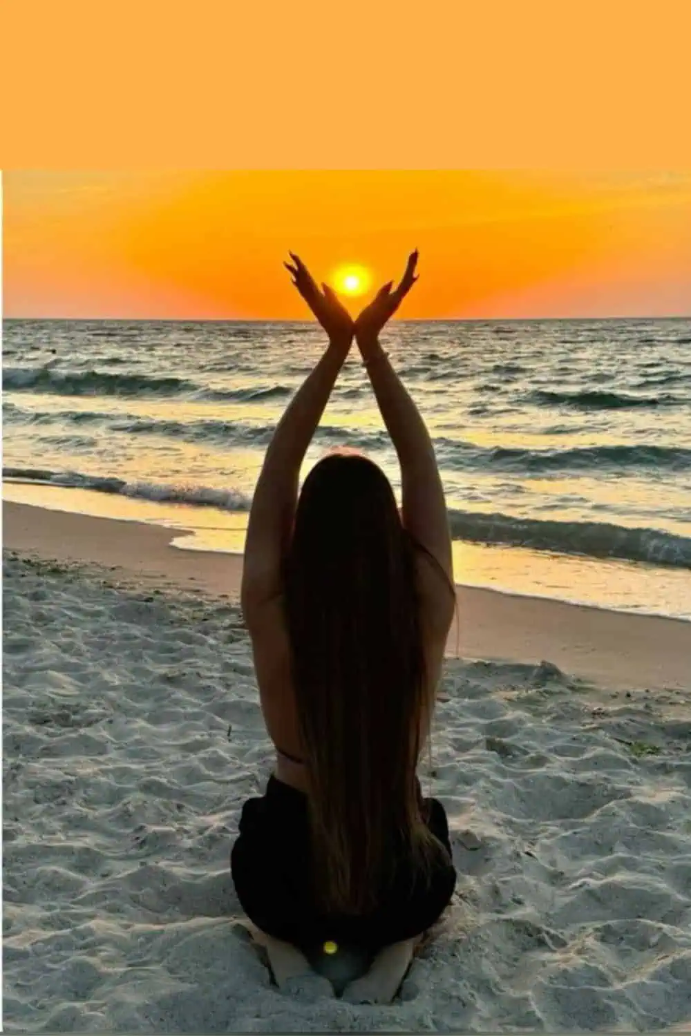 Girl kneeling on the beach at sunset with arms raised, cupping the sun in her hands. Aesthetic, backlit summer photo pose with golden hour lighting, ocean waves, and soft sand.