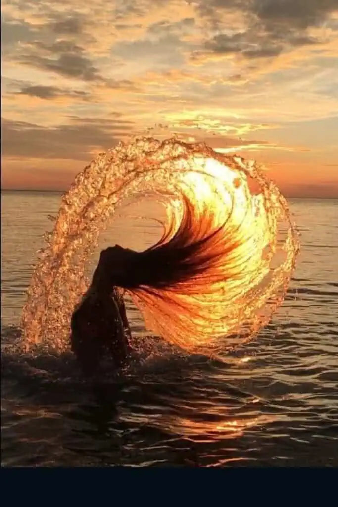 Woman flipping her wet hair in a dramatic arc at sunset, creating a glowing water splash shaped like a fountain—perfect summer mermaid pose captured against a golden sky.