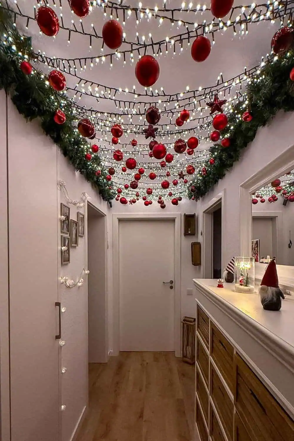 Hallway ceiling decorated for Christmas with rows of twinkling white lights, hanging red ornaments, and green garland along the edges for a festive and immersive holiday look.