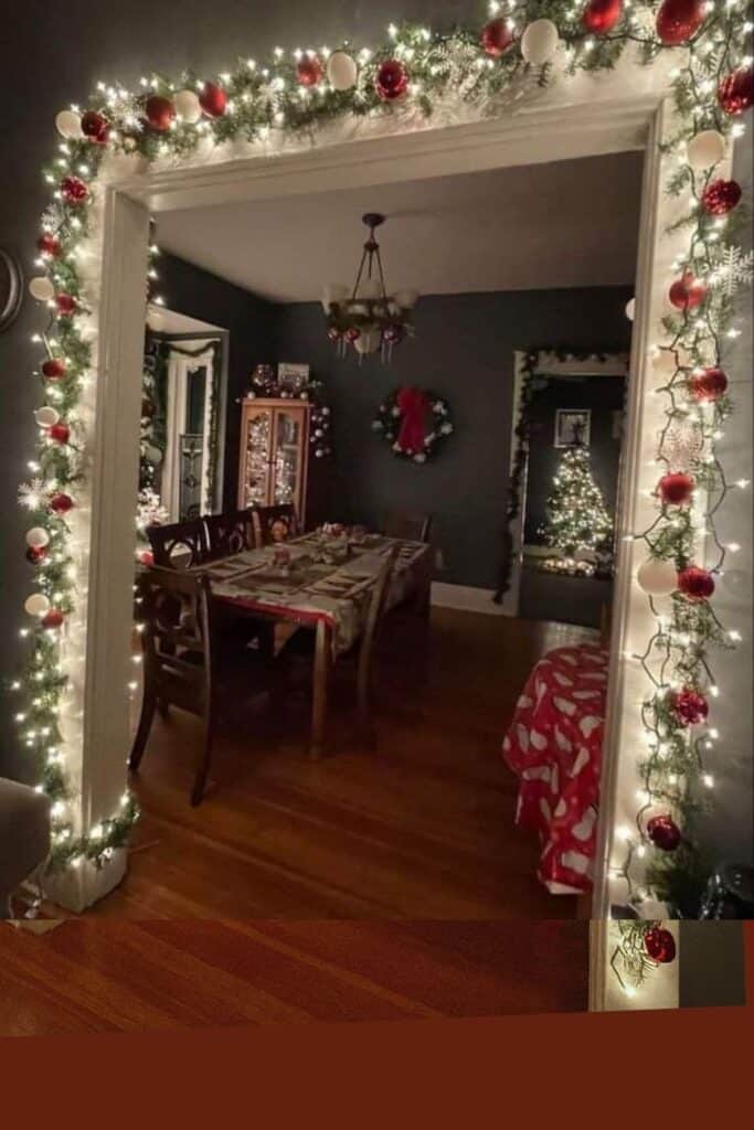 Cozy Christmas dining room decorated with glowing garland, red and white ornaments, twinkling string lights, a holiday dining table, and a Christmas tree in the background.