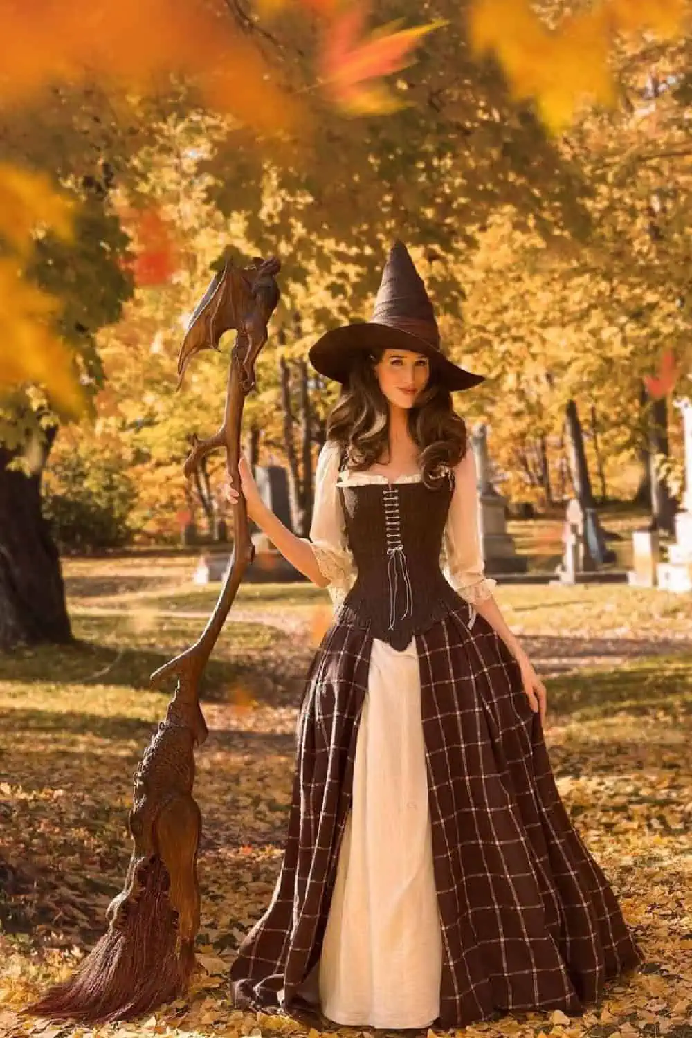 Woman in a classic witch costume with a brown plaid skirt, cream blouse, corset top, and pointed hat, holding a dragon-shaped broomstick in a fall forest with golden leaves.