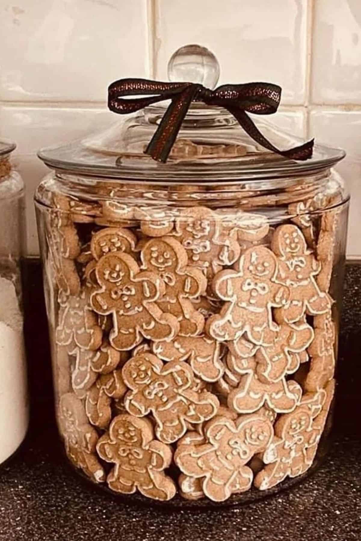 Glass jar filled with gingerbread cookies and topped with a black ribbon, sitting on a kitchen counter next to jars of flour and sugar; cozy and festive Christmas kitchen decor idea.