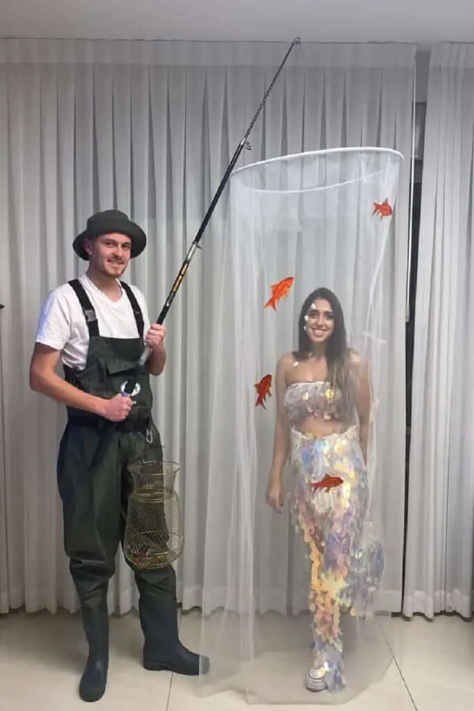 Funny couples Halloween costume: a fisherman in green waders and boots holding a fishing rod and basket stands next to his “biggest catch,” a woman inside a tall tulle net wearing iridescent sequin “fish scale” outfit with orange goldfish decorations.