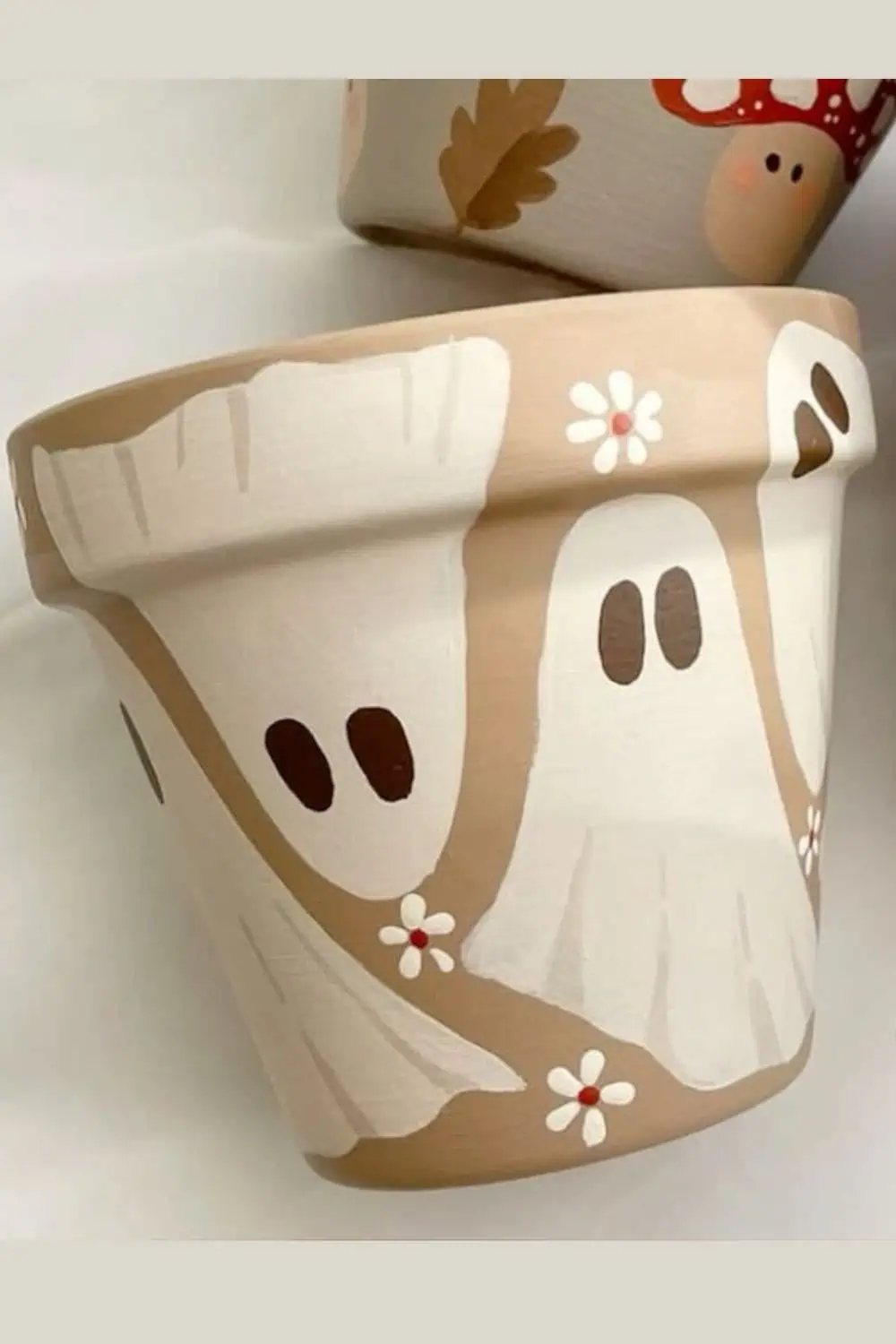 Hand-painted terracotta pot decorated with minimalist white ghosts and daisy flowers on a beige background, perfect for early fall or Halloween decor.
