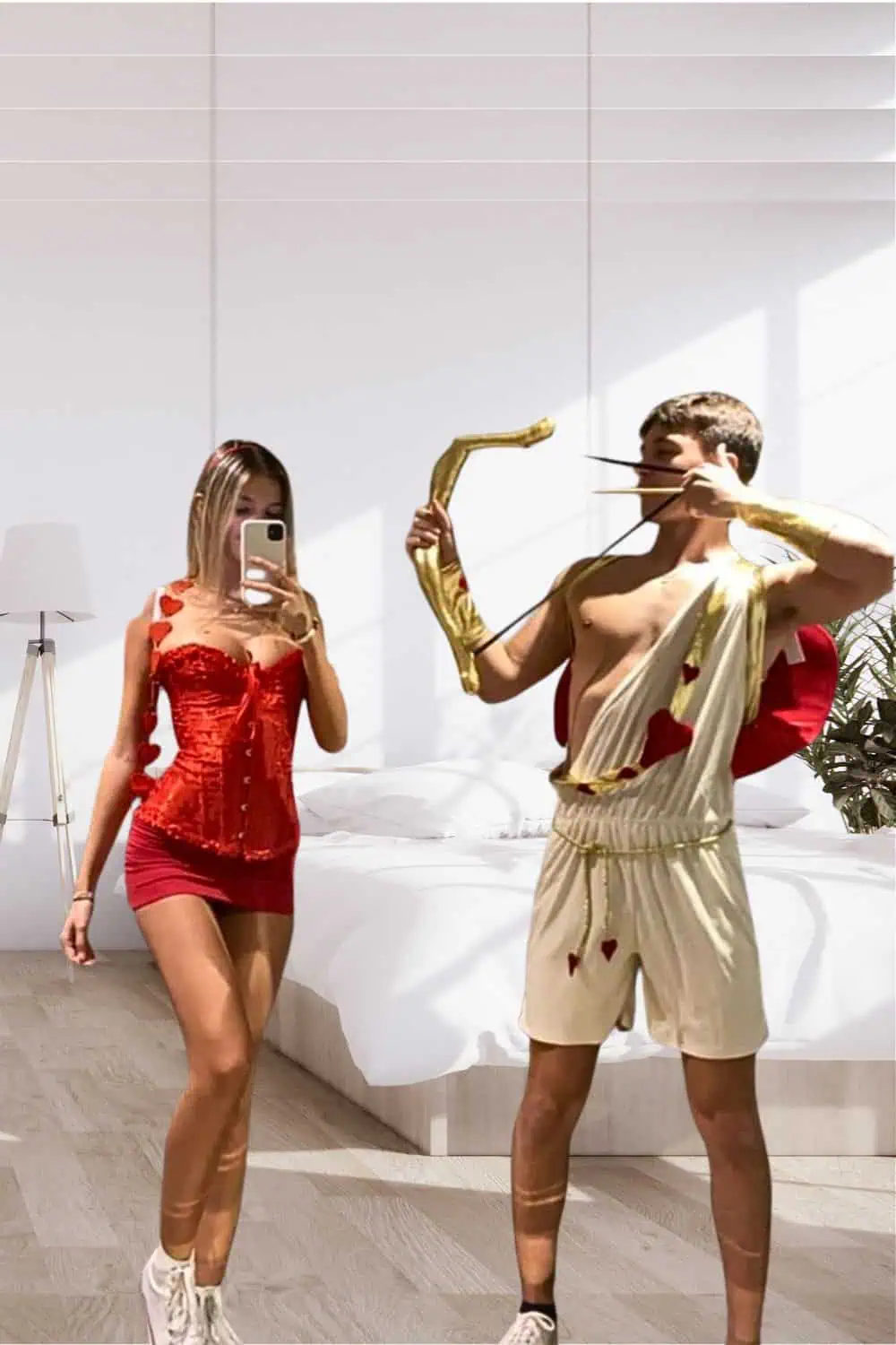 A young couple dressed in matching Cupid-inspired Halloween costumes poses in a modern bedroom. The woman wears a red corset and mini skirt with heart details and angel wings, while the man wears a white toga-style outfit with red heart accents, gold trim, and holds a golden bow and arrow. This fun and flirty costume idea is perfect for couples looking for creative, romantic Halloween looks.
