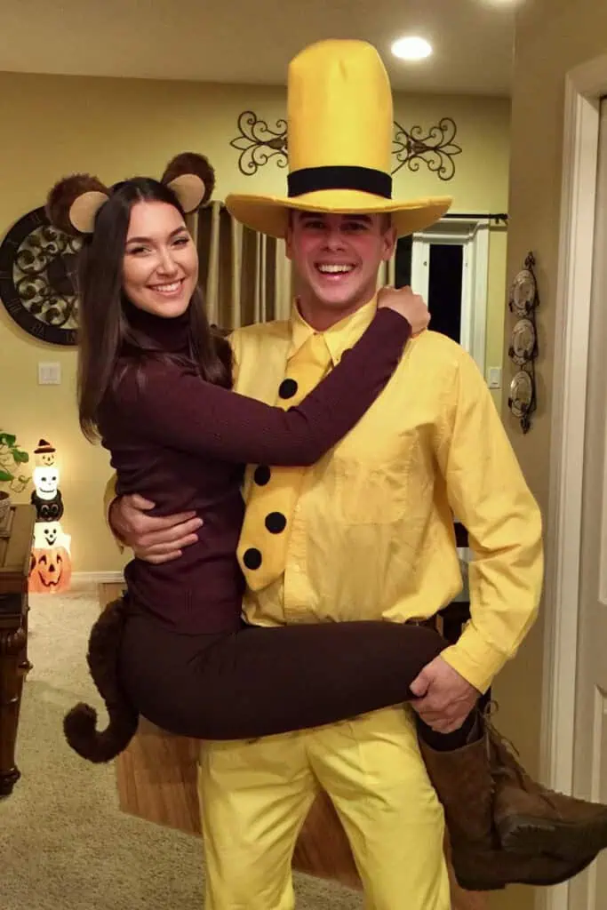 Couple wearing Curious George Halloween costumes with the woman dressed as George the monkey in brown outfit with ears and tail, and the man dressed as the Man in the Yellow Hat with a tall yellow hat, yellow shirt, pants, and boots.
