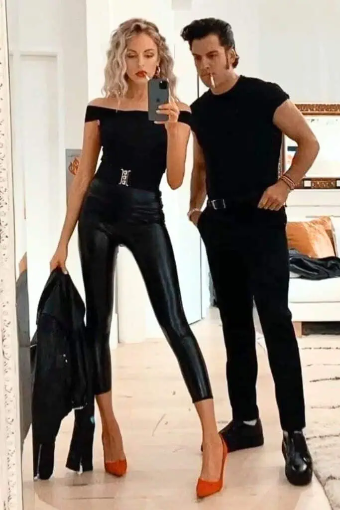 Couple dressed as Sandy and Danny from Grease in matching black outfits, red heels, leather jacket, and slicked-back hair, posing confidently indoors for a stylish DIY Halloween couples costume photo for high school, college, or adults.