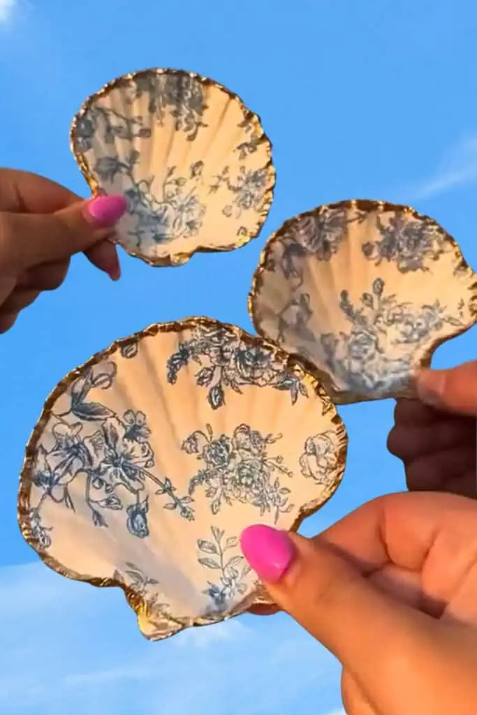 Three decorative jewelry dishes made from white scallop seashells lined with blue floral napkins and finished with gold-painted edges, creating an elegant coastal-style flat lay.