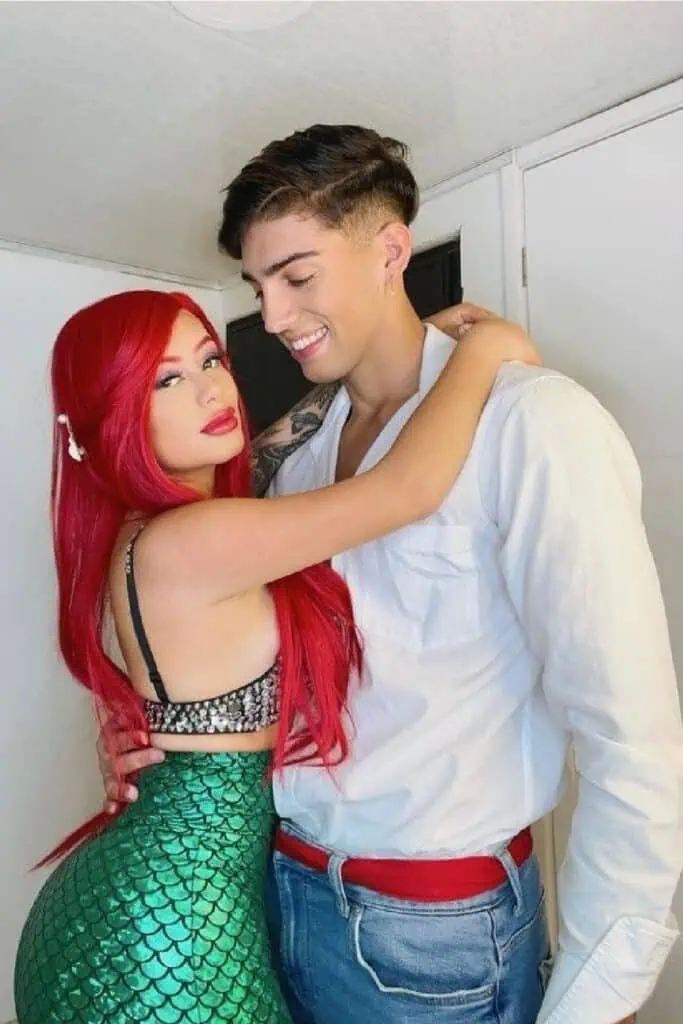 Couple dressed in a Disney Little Mermaid inspired Halloween costume. The woman is styled as Ariel with a long red wig, seashell-inspired top, and shiny green mermaid skirt. The man wears a white shirt, jeans, and a red belt, resembling Prince Eric. Cute DIY couples costume idea for Halloween or cosplay.