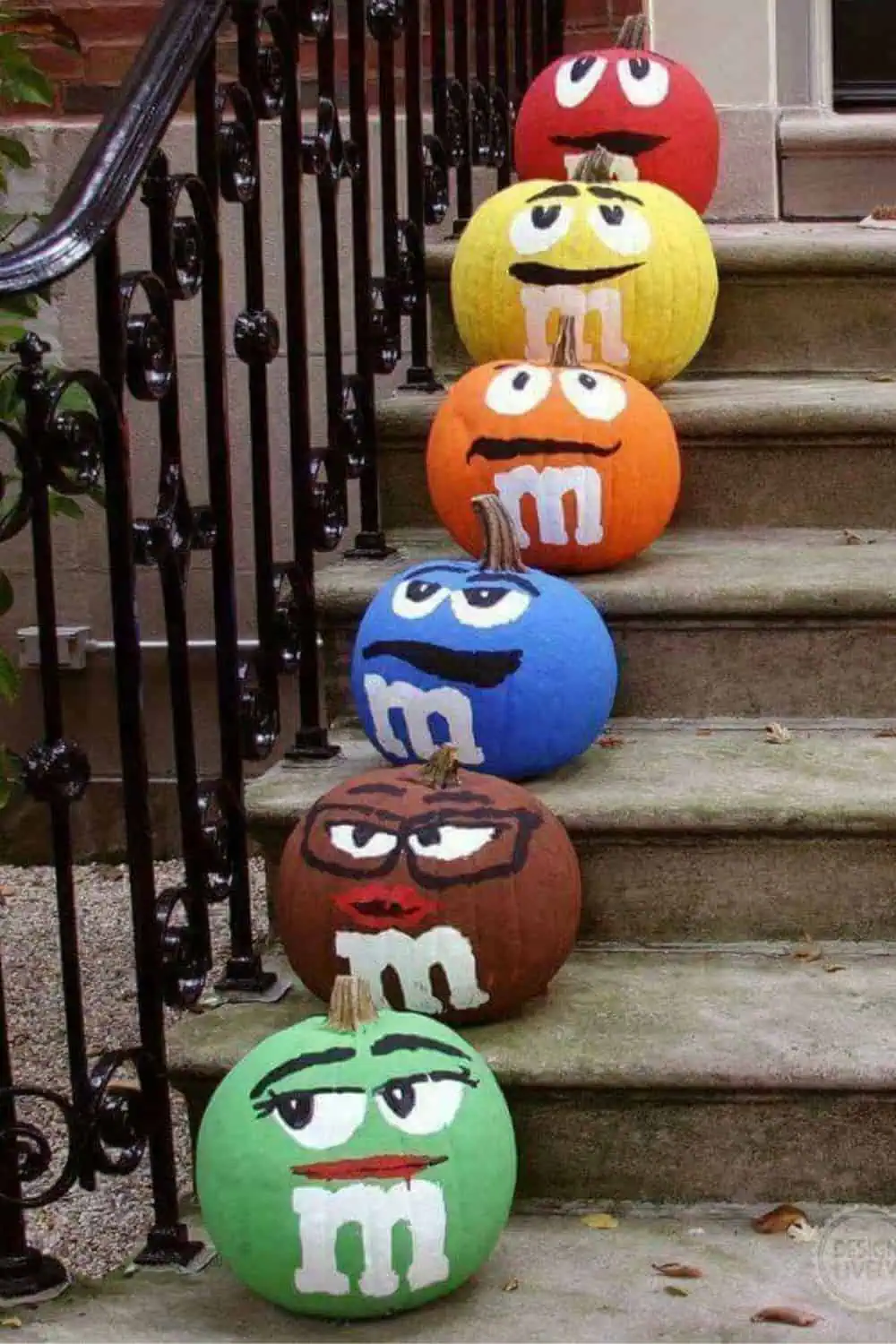Colorful M&M-themed pumpkins painted in red, yellow, orange, blue, brown, and green arranged on outdoor stairs as a fun no-carve Halloween decoration for 2025.”