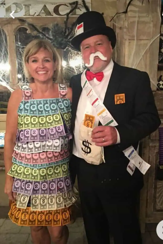 Funny Monopoly couples Halloween costume: woman in a tiered dress covered with play money, man as Mr. Monopoly in a suit with top hat, white mustache, deed‑card sash, and a money bag.