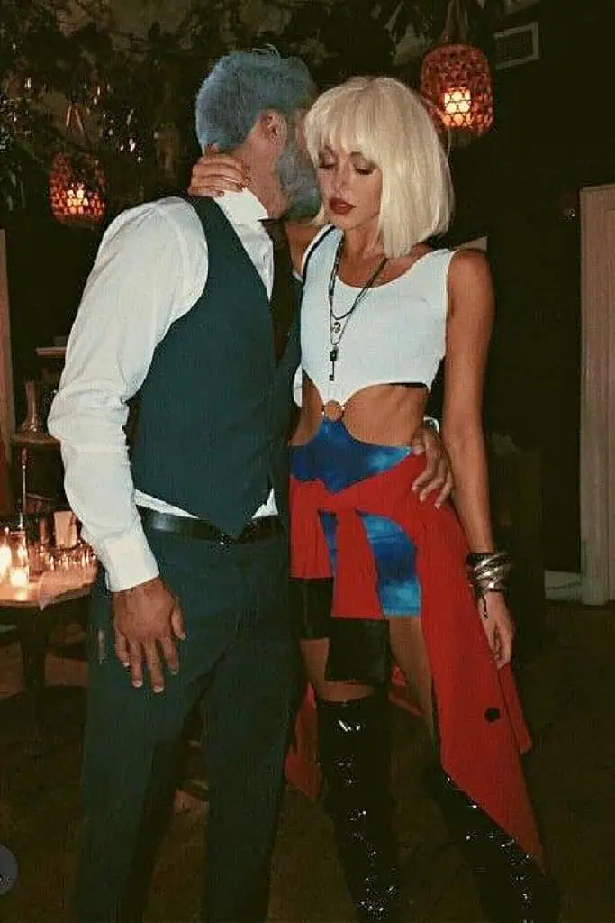 A couple dressed in coordinating Halloween costumes inspired by the movie Pretty Woman. The woman wears a platinum blonde wig, white cut-out tank top, blue mini skirt, red jacket, and black thigh-high boots. The man is dressed in a sharp navy suit with a white shirt and vest, resembling the business look of Edward Lewis. The photo is taken indoors with moody lighting and candles in the background, perfect for a romantic or movie-themed costume party.