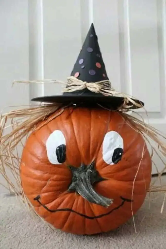 DIY painted pumpkin decorated as a witch with hand-painted cartoon eyes, a smile, raffia hair, and a black witch hat with colorful polka dots. Cute no-carve Halloween pumpkin idea for kids and fall decorating.