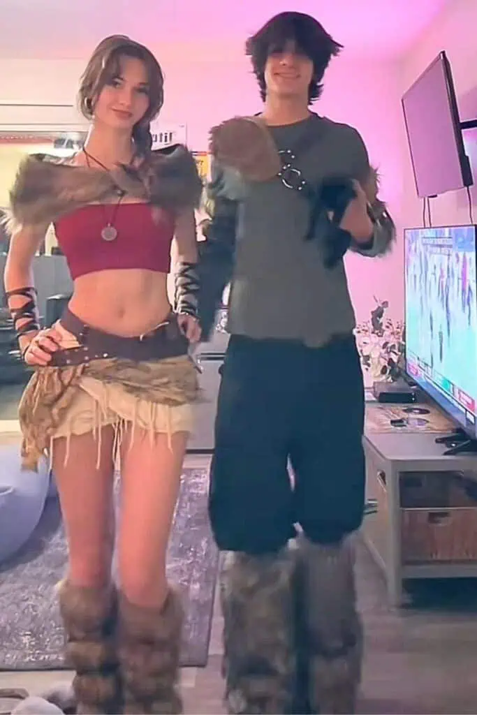 Couple dressed as Astrid and Hiccup from How to Train Your Dragon in DIY Halloween costumes, wearing fur-lined boots and Viking-inspired outfits, standing indoors under soft pink lighting.
