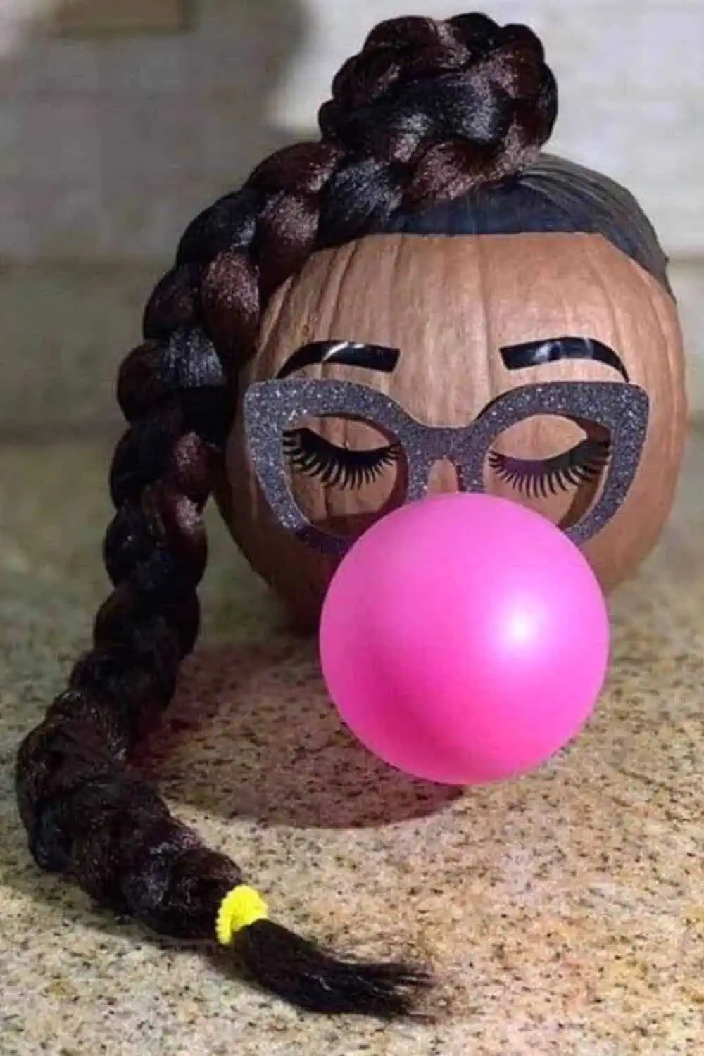 Funny no-carve pumpkin decorated as a girl blowing pink bubblegum, with braided hair, glitter glasses, and painted eyelashes — a creative pumpkin painting idea for 2025.