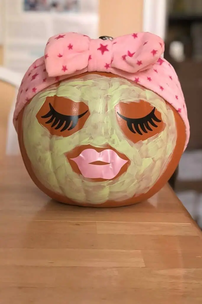 Pumpkin painted with a green face mask, long lashes, pink lips, and a pink spa headband — a cute no-carve Halloween pumpkin decoration.