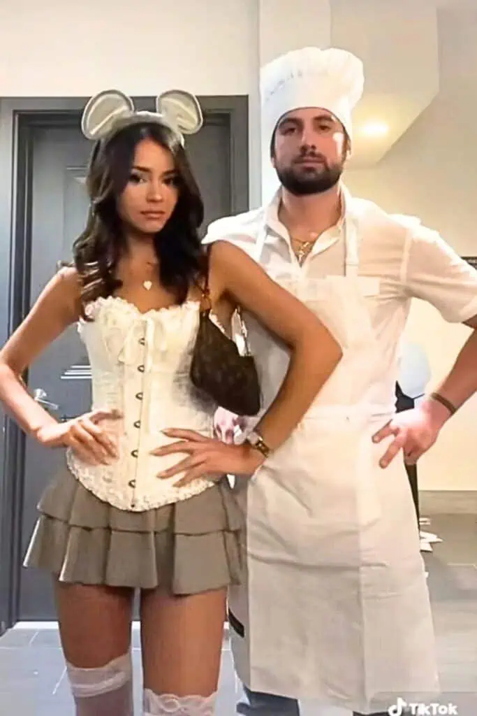 Couple dressed as Remy and Linguini from Ratatouille for Halloween, with the woman wearing a white corset and mouse ears and the man in a chef’s outfit and apron, posing indoors in a trendy DIY couples costume cute.