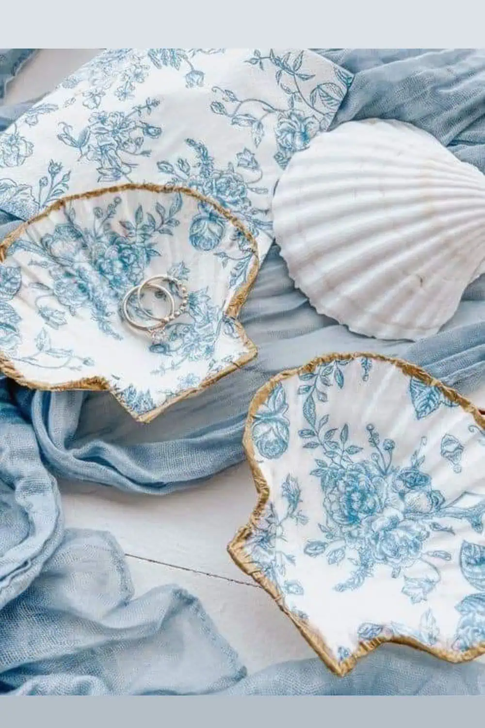 Easy DIY seashell jewelry dishes made with Dollar Store supplies, featuring blue floral napkin decoupage and gold-painted edges, styled with rings and soft fabric.