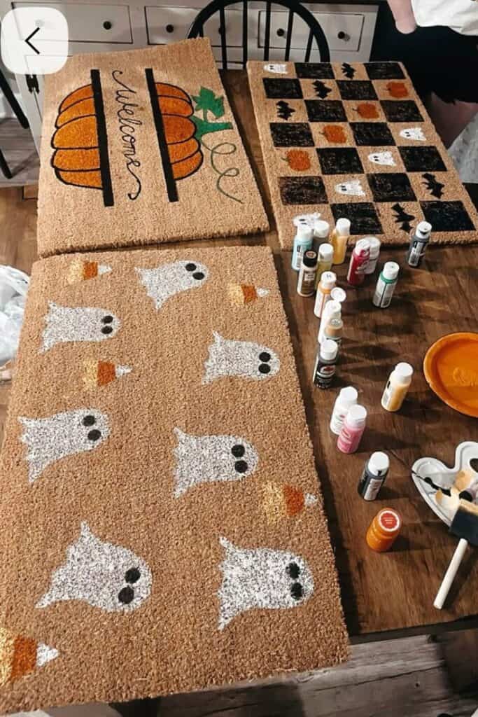DIY Halloween doormat craft made with coir mats, dollar store paint, and stencils featuring designs like pumpkins, ghosts, candy corn, and bats, painted on a table with brushes and supplies.
