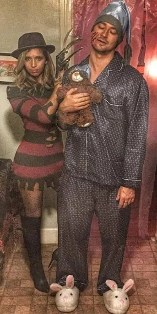 Couple in a funny Halloween costume with woman dressed as Freddy Krueger in a striped dress and claw glove, and man dressed as a sleepy pajama victim holding a teddy bear with claw marks and slippers.