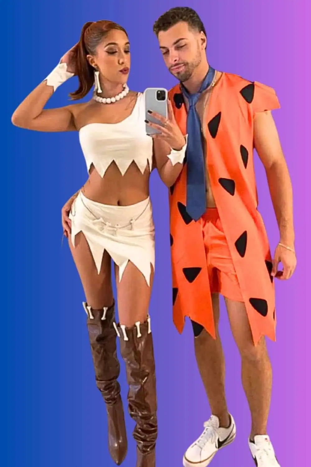 Couple dressed as Fred and Wilma Flintstone for Halloween. Wilma wears a white crop top and skirt with pearl accessories and tall brown boots, while Fred wears an orange tunic with black spots, matching shorts, a blue tie, and sneakers.