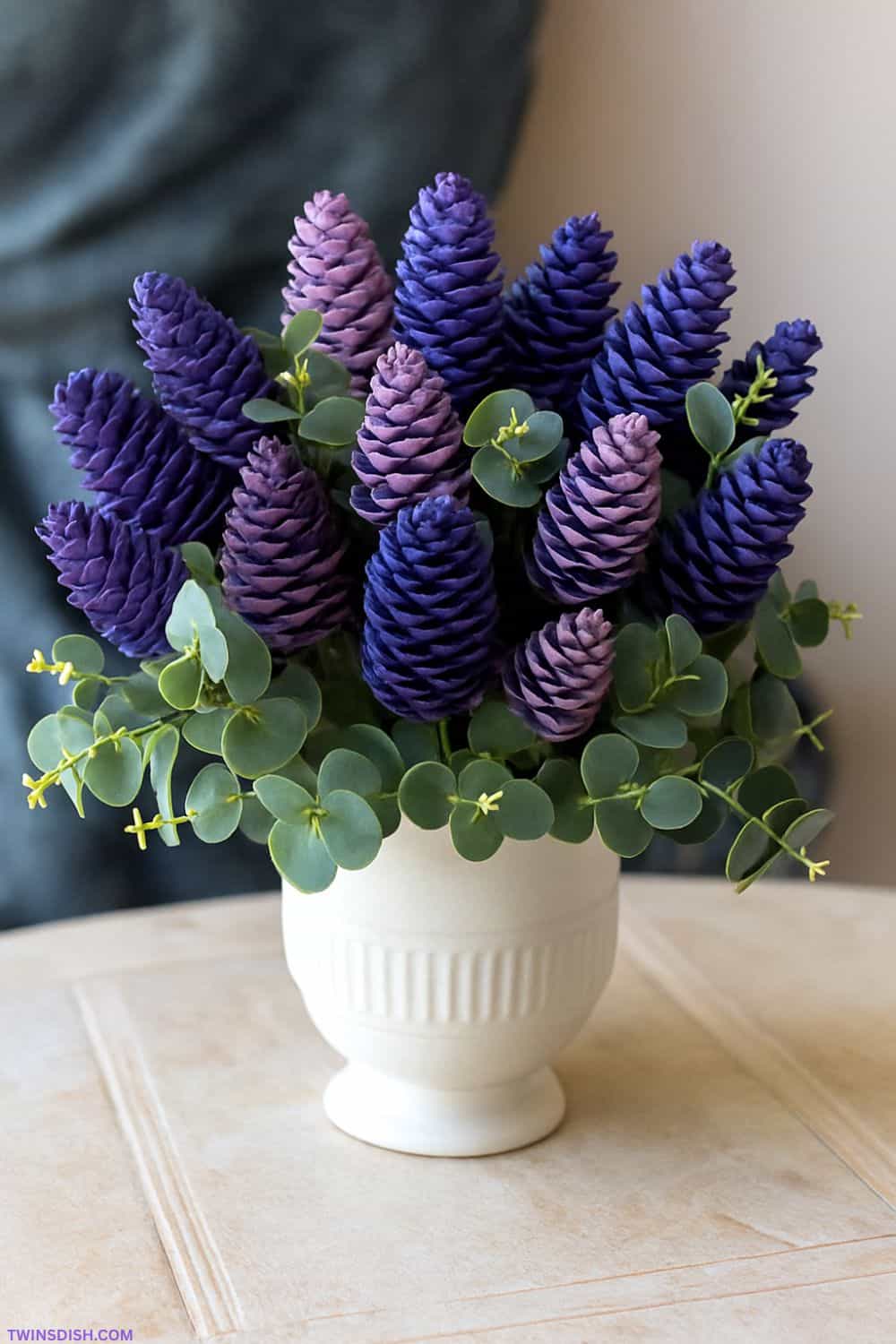 DIY bouquet of pinecones painted to look like lilacs in shades of purple and lavender, arranged with eucalyptus leaves in a white ceramic vase on a neutral background.