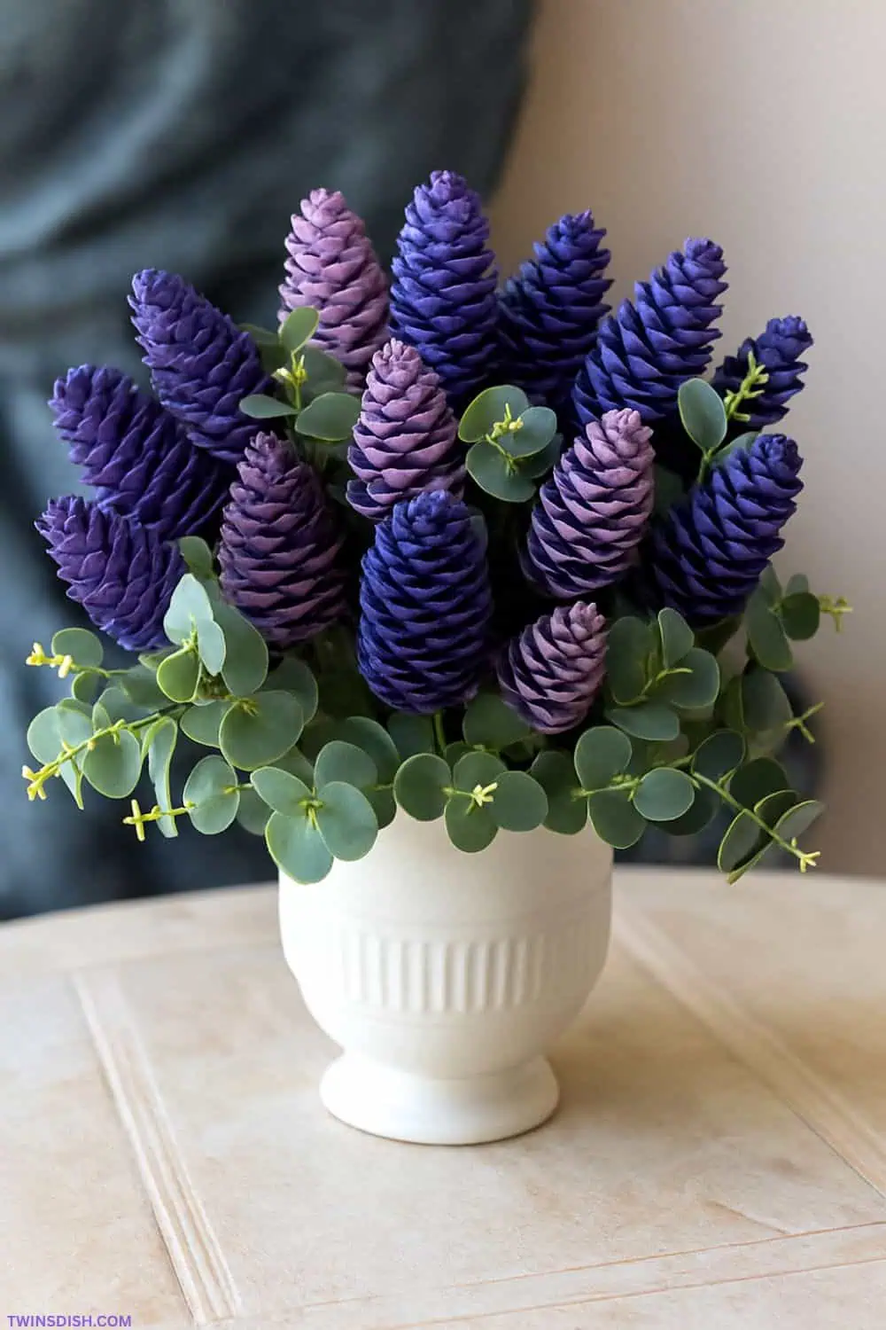 DIY bouquet of pinecones painted to look like lilacs in shades of purple and lavender, arranged with eucalyptus leaves in a white ceramic vase on a neutral background.