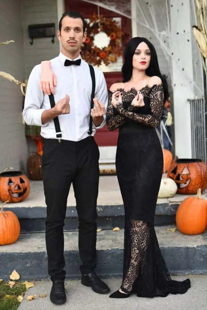 Couple dressed as Morticia and Gomez Addams in a black lace gown and formal suspenders, posing on a Halloween-decorated porch with pumpkins and spider webs.