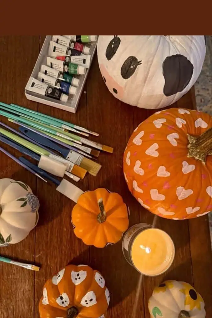 DIY no-carve painted pumpkins decorated with cute designs including hearts, ghosts, sunflowers, roses, and a cow face. Pumpkins are displayed on a wooden table with paintbrushes, acrylic paints, and a candle.
