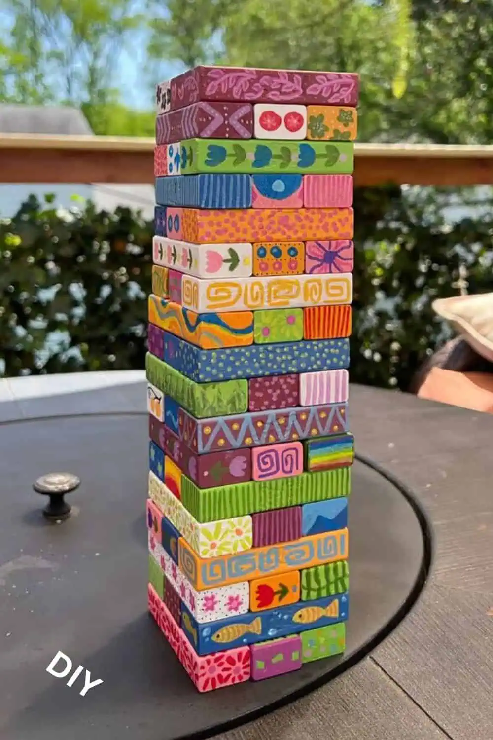 Colorful hand-painted Jenga blocks stacked into a tower with detailed patterns, flowers, and designs, displayed outdoors as a fun DIY summer craft for kids and adults.