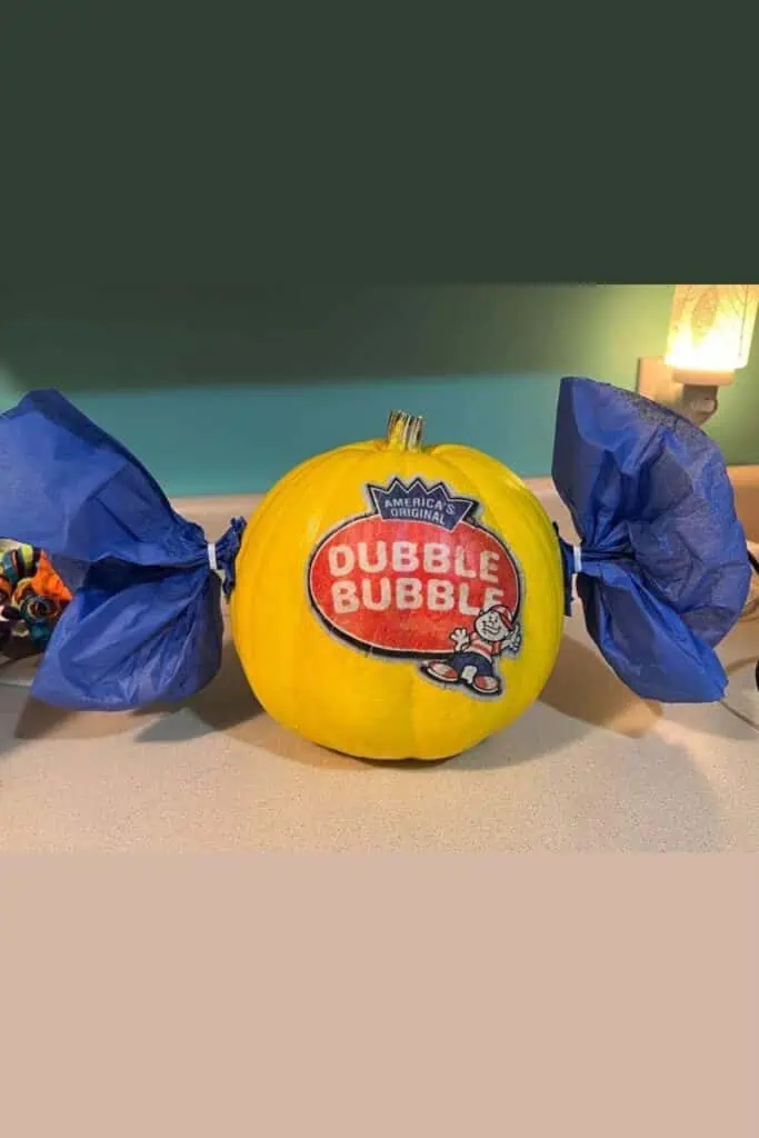 No-carve painted pumpkin decorated to look like a Dubble Bubble gum piece, featuring a bright yellow pumpkin with the Dubble Bubble logo on the front and blue tissue paper “twists” on each side to resemble candy wrappers.