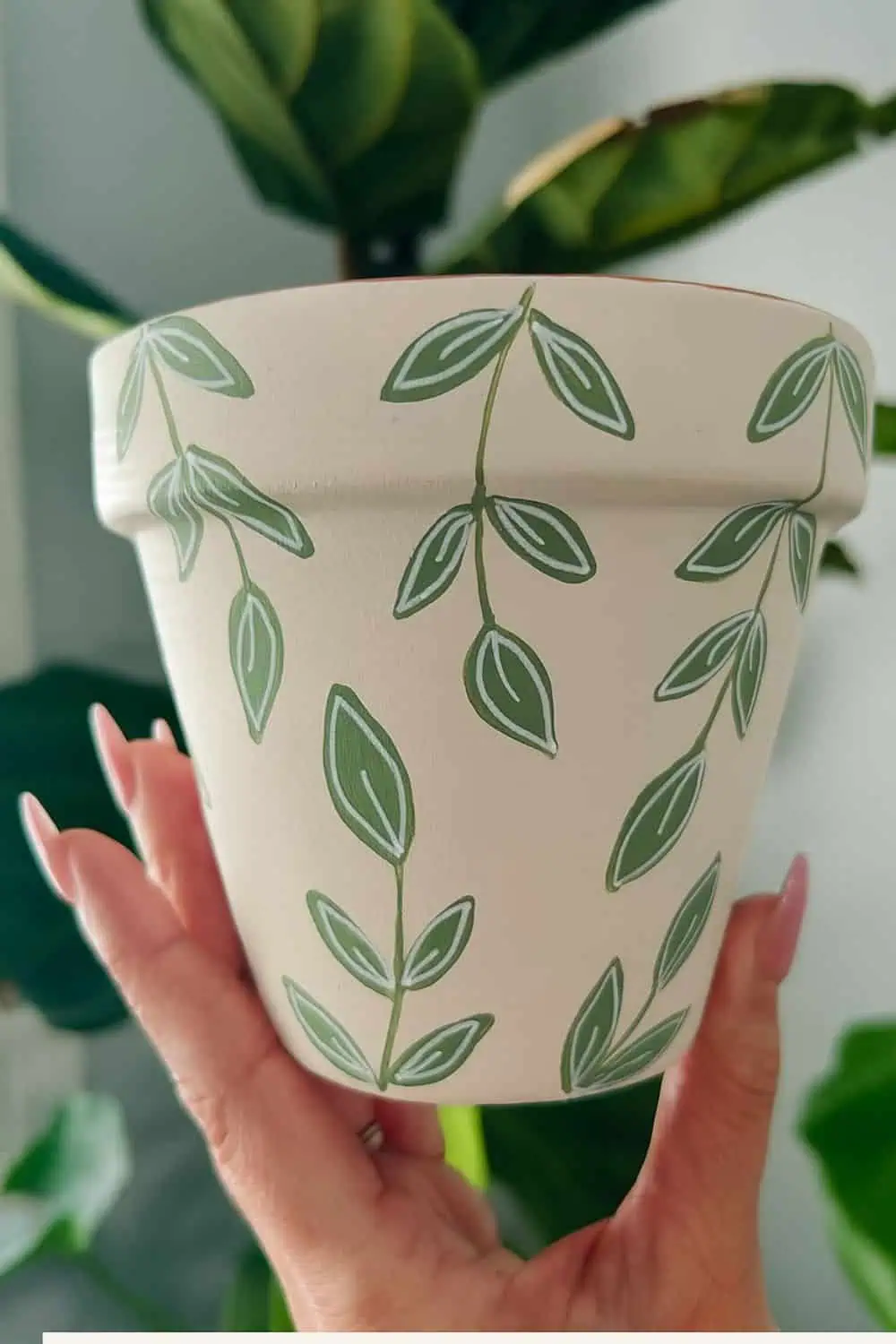 Hand-painted terracotta planter featuring delicate green vine and leaf designs held up by a hand with long pink nails, with a soft indoor plant background.
