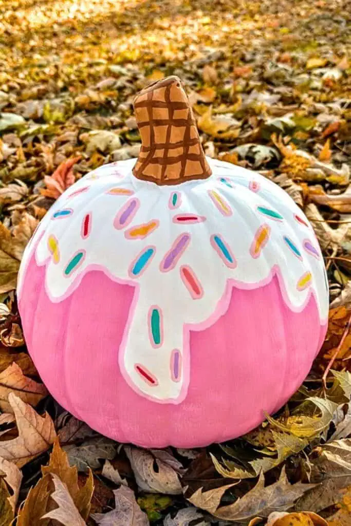 Pink pumpkin painted to look like an ice cream cone with white dripping “frosting,” rainbow sprinkles, and a waffle cone stem — a cute and creative no-carve Halloween pumpkin idea.