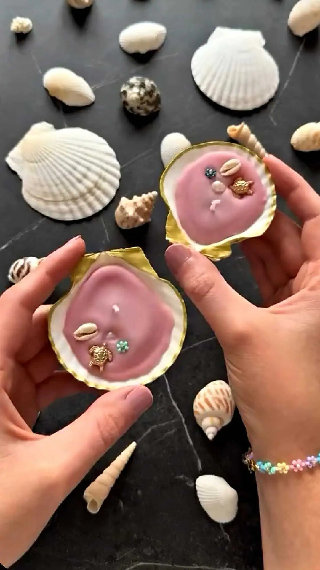 Two handmade seashell tealight candles with pink wax, gold trim, and tiny beach-themed embellishments, held above a dark surface surrounded by assorted sea shells.