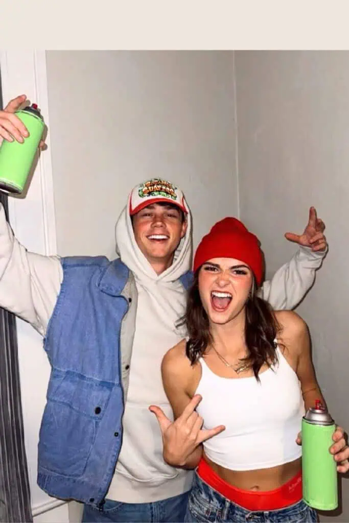 Couple dressed as Subway Surfers characters for Halloween costumes for teens and College students, wearing a red beanie, white crop top, hoodie, and denim, each holding green spray paint props while posing playfully indoors.