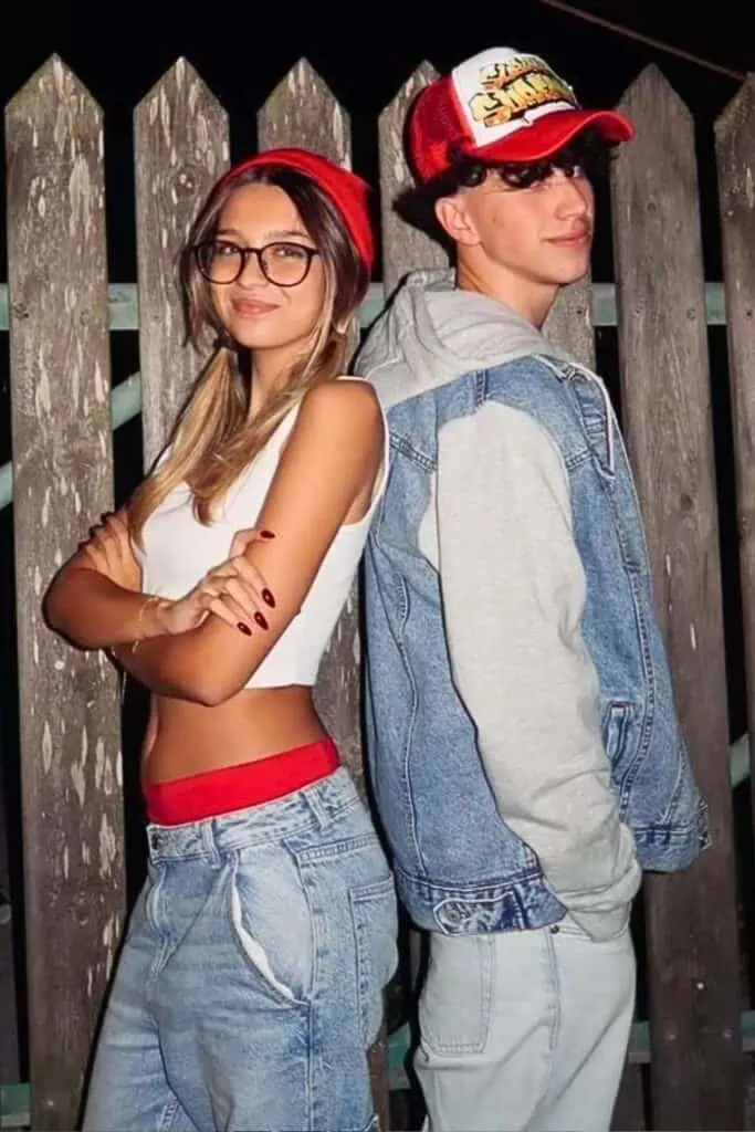 Couple wearing DIY Subway Surfers Halloween costumes, standing back to back in denim outfits, with a red beanie and Subway Surfers hat, posing against a wooden fence at night.