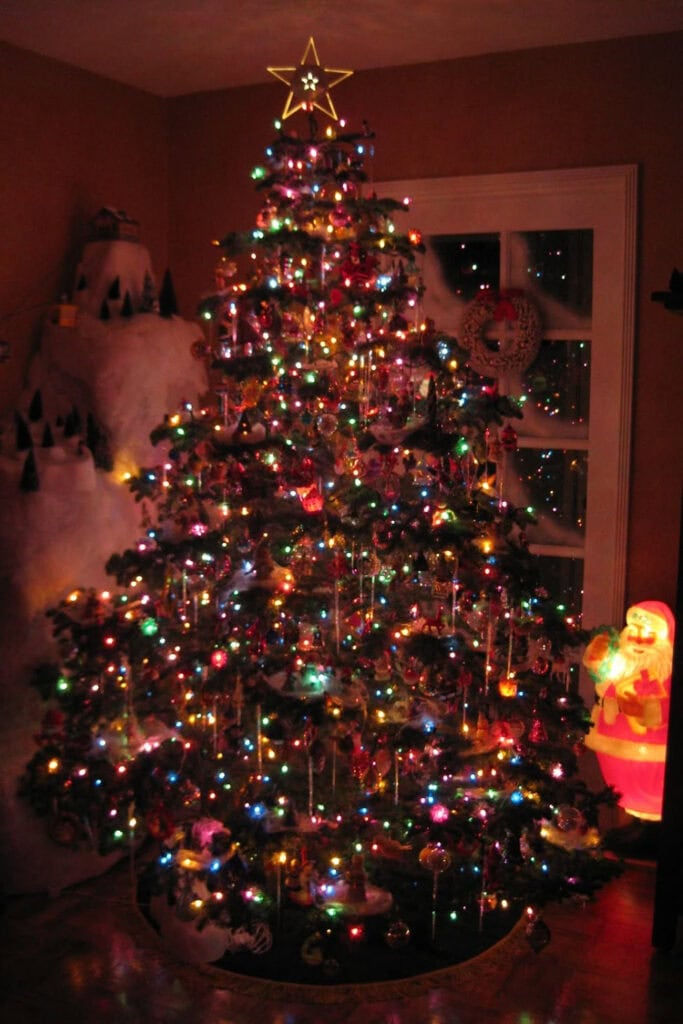 Vintage 1980s–1990s Christmas tree decorated with colorful lights, glass ornaments, tinsel, and a glowing star topper, surrounded by nostalgic holiday decorations including a light-up Santa Claus figure.