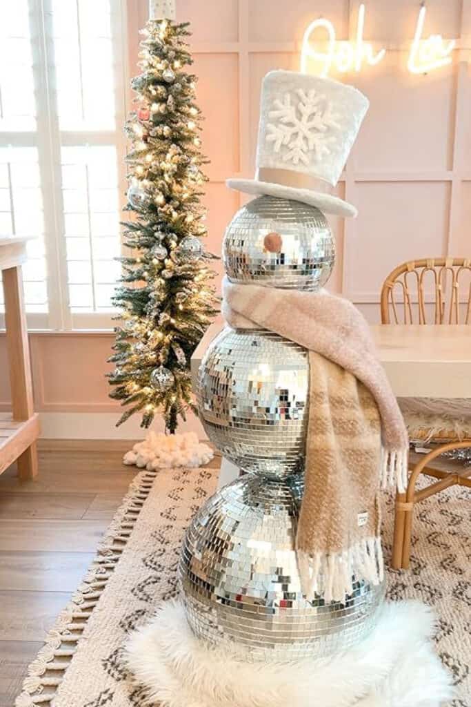 A snowman made of stacked mirrored disco balls, topped with a fuzzy white top hat and wrapped in a neutral beige plaid scarf. It stands on a white faux fur rug in a pastel pink room next to a slim, flocked Christmas tree with silver ornaments. This image reflects the Retro and Vintage Christmas decor trend for 2025, combining 1970s disco style with cozy, modern holiday flair.