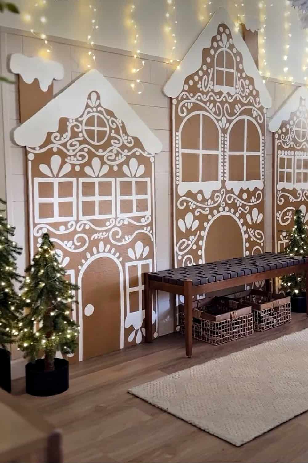DIY gingerbread house Christmas wall decor made from cardboard with white painted details. Oversized gingerbread house decorations with string lights and mini Christmas trees.