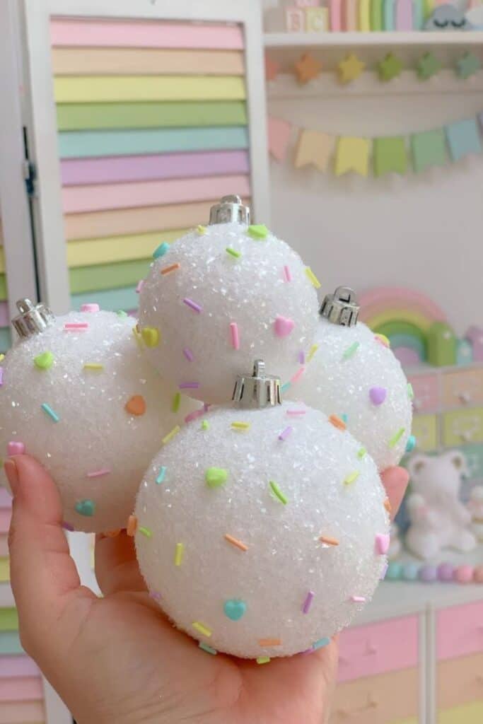 Hand holding pastel candy sprinkle Christmas ornaments, featuring white glitter balls decorated with colorful pastel heart and sprinkle accents, perfect for whimsical kids’ themes like Candy Land, gingerbread, and playful holiday decor.