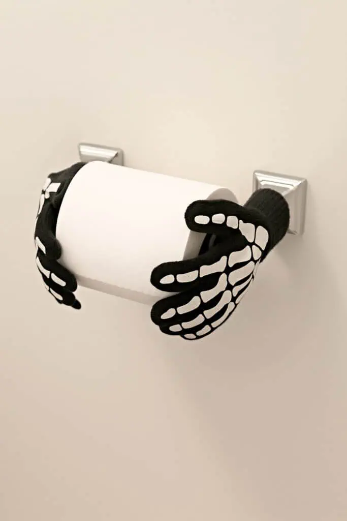 Halloween-themed toilet paper holder with black skeleton gloves gripping a white toilet paper roll on a chrome wall mount against a light beige wall.