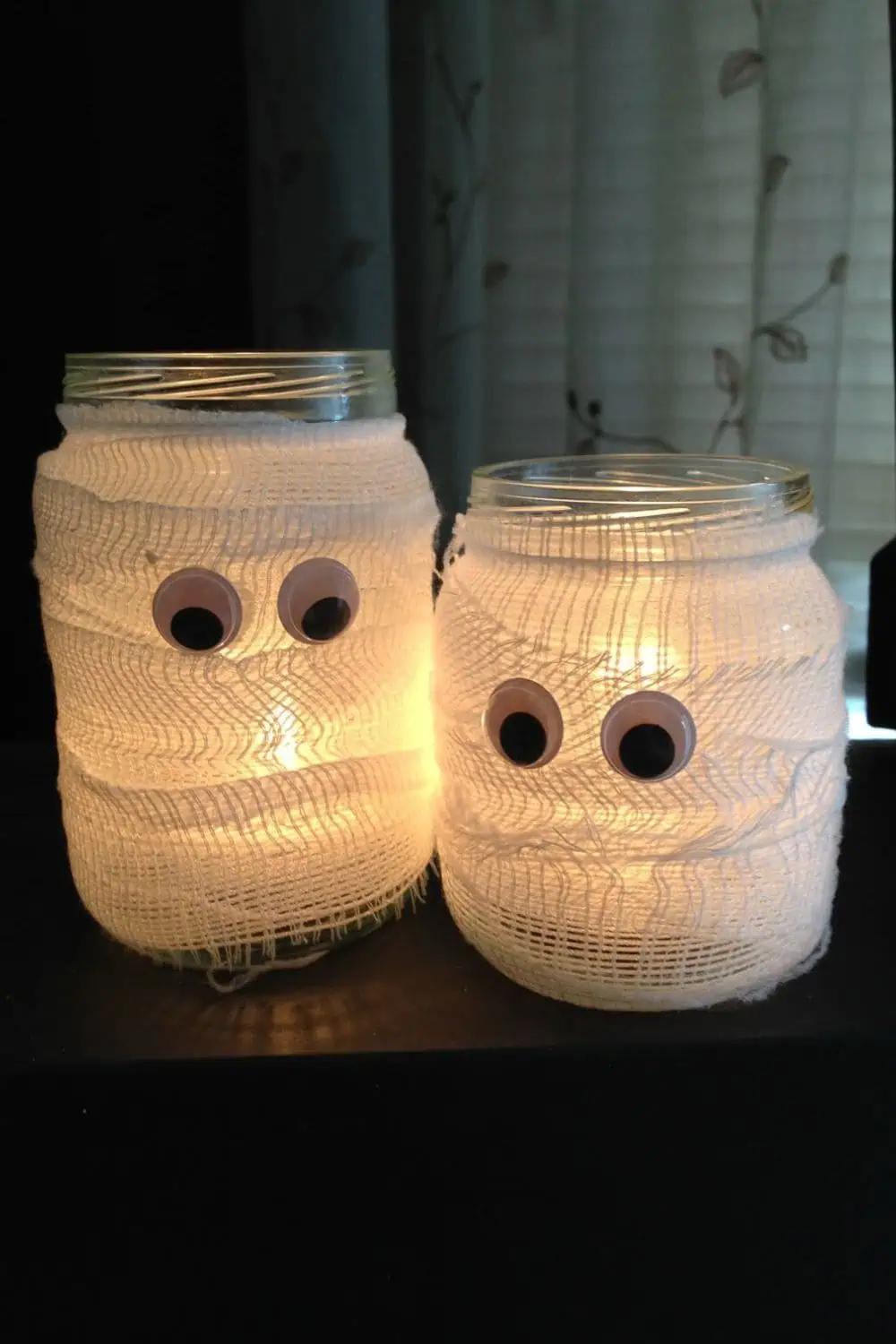 Two glowing mason jar mummy lanterns wrapped in white gauze with googly eyes—simple dollar store Halloween DIY using LED tea lights. Kid friendly craft with supervision.