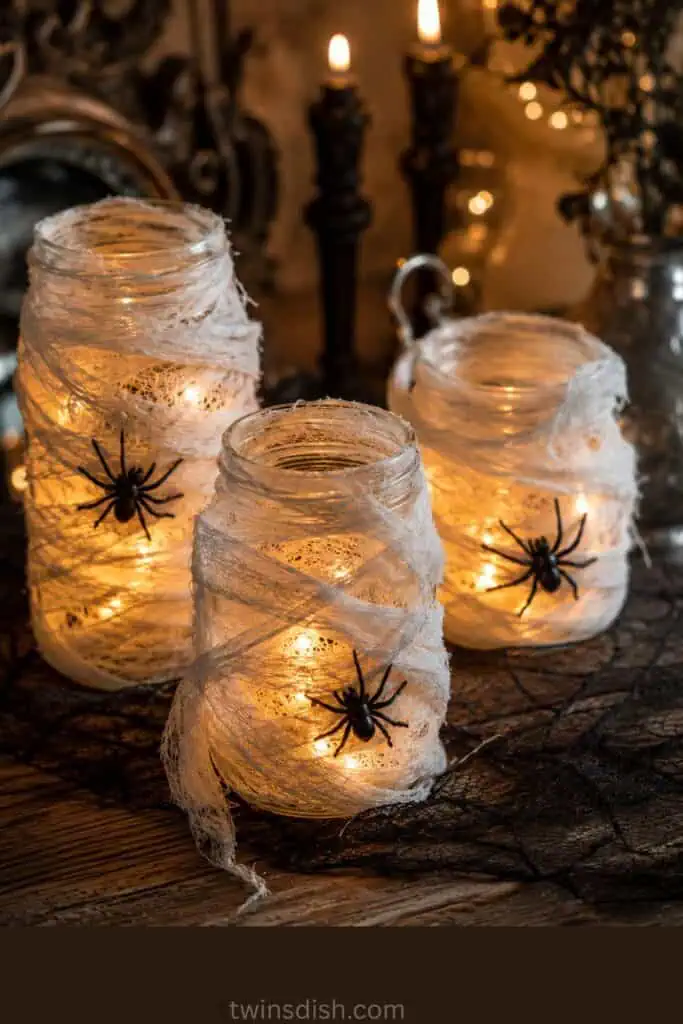 Three mason jars wrapped unevenly in white cheesecloth with black spiders and warm glowing lights for Halloween.
