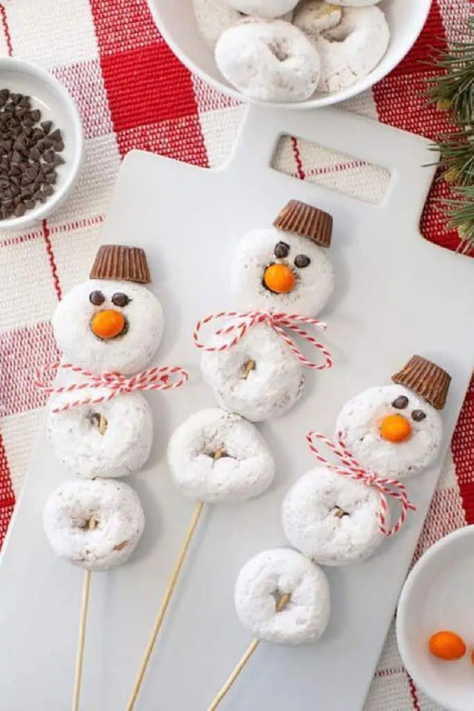 Snowman donut skewers made with powdered sugar donuts, chocolate hats, candy noses, and twine scarves arranged on a cutting board for a festive Christmas treat.