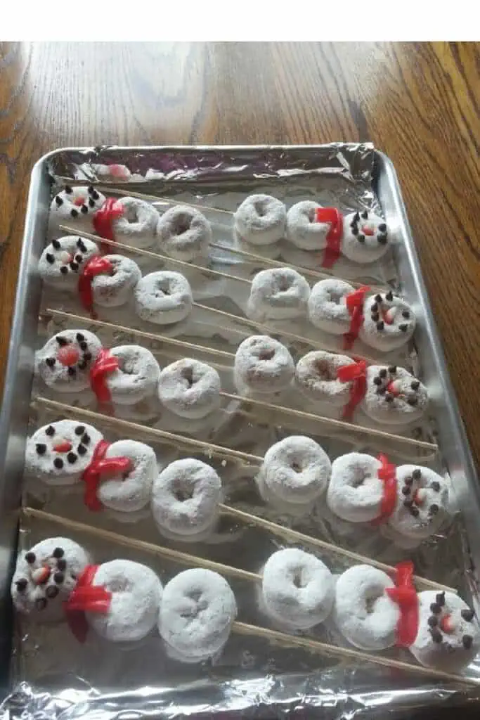 Snowman donut skewers made with powdered sugar donuts, candy faces, and red candy scarves arranged on a baking tray for a festive Christmas treat.